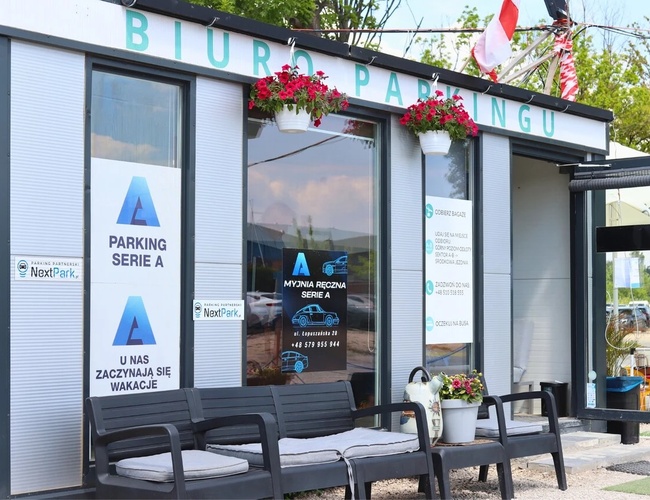 Najlepšia parkovacia séria A – Parkovacie pracovisko s dekoratívnymi kvetmi. Pred budovou stoja lavičky s vankúšmi. Okolo rastú stromy.