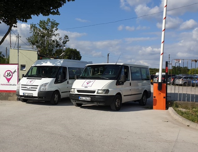 JG Parking - Dos furgonetas blancas están paradas junto a la barrera. El aparcamiento está cercado con una valla metálica. Al fondo se pueden ver coches aparcados.