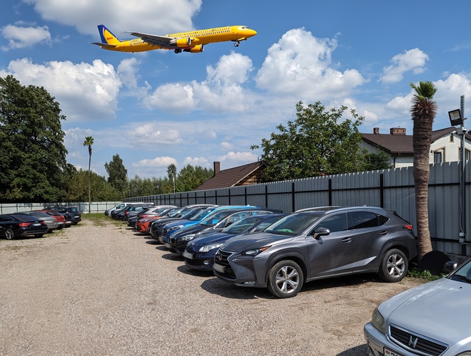 Aparcamiento P60 - Los coches están en una superficie de grava. Un avión amarillo vuela sobre el aparcamiento. El área está cerrada con una valla de metal.