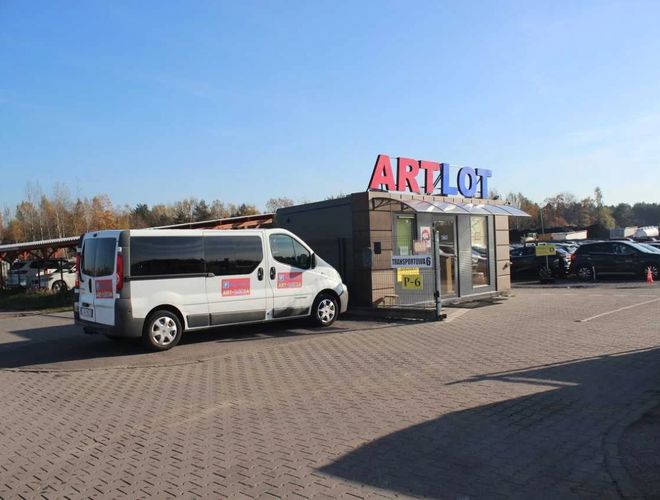 ArtLot 2 - Minibus stoi przed biurem na parkingu. Teren jest utwardzony. W tle widać zadaszone miejsca postojowe.