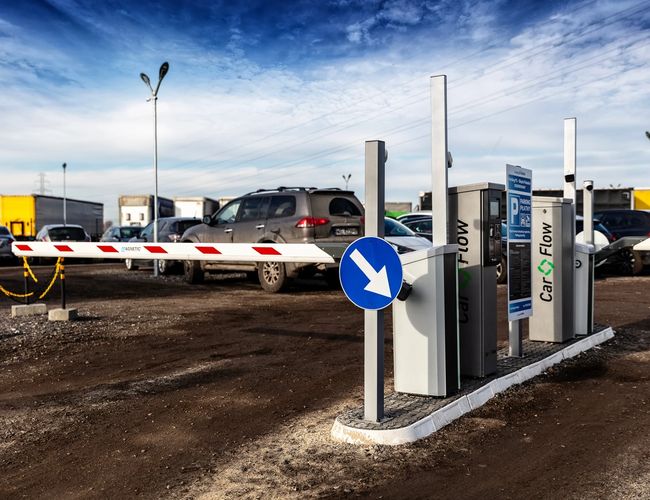 Parking Okęcie Południe – Wjazd z szlabanem i oznaczeniami drogowymi. Teren jest żwirowy z kilkoma zaparkowanymi samochodami.