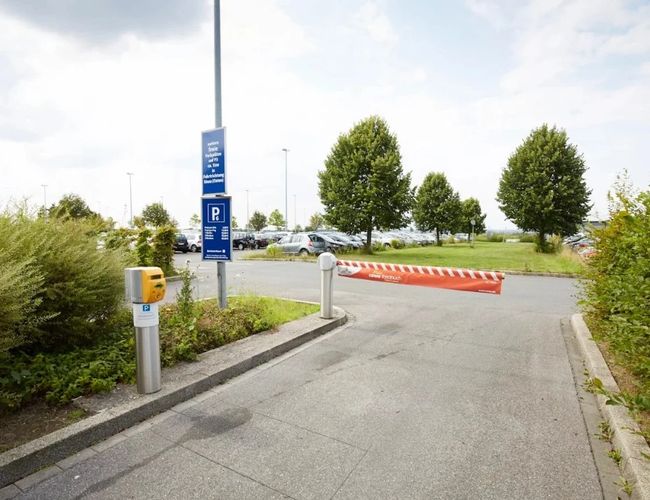Droga prowadząca do parkingu z automatycznym szlabanem. Po lewej stronie znajdują się znaki informacyjne, a w tle widoczny jest teren parkingu otoczony zielenią.