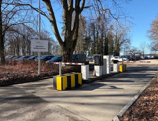 Wjazd na teren parkingu z automatycznymi szlabanami oraz urządzeniami do kontroli wjazdu. Po obu stronach znajdują się drzewa.
