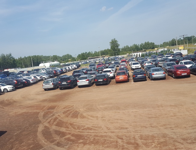 AAP-66 - Parking jest otwarty i nieutwardzony. Na placu stoi wiele samochodów. W tle widać zieleń i ogrodzenie.