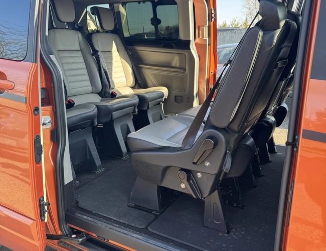 Estacionamiento Wings - Puertas abiertas del autobús naranja. El interior tiene tres filas de asientos grises. El aire acondicionado es visible en el techo.