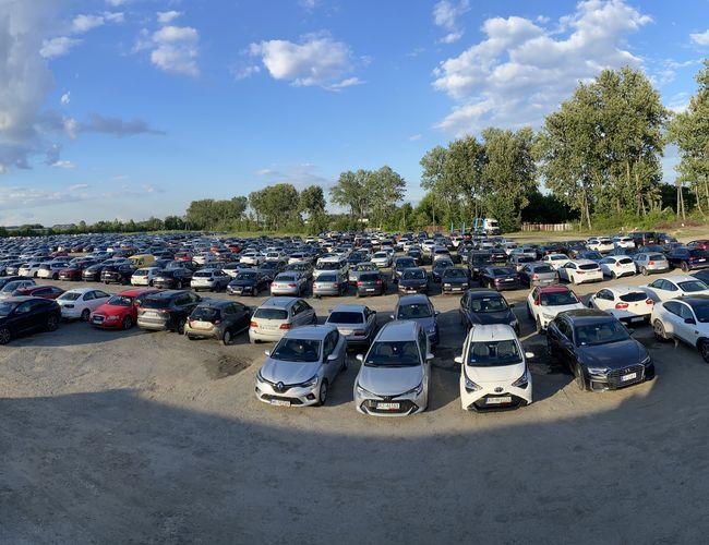 Odkryj-Parking - Widoczny jest duży, otwarty parking pełen samochodów. W tle znajdują się drzewa i jasne niebo. Teren jest nieogrodzony.