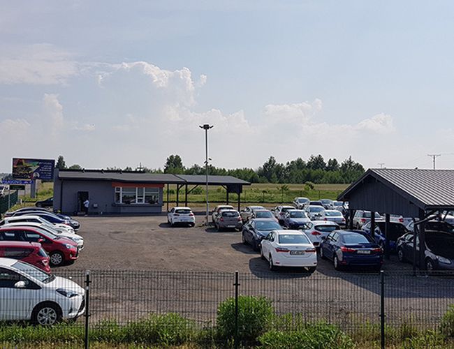 Parking MTM – plac z samochodami otoczony jest ogrodzeniem. Teren jest utwardzony i dobrze oświetlony. W tle widać budynek.