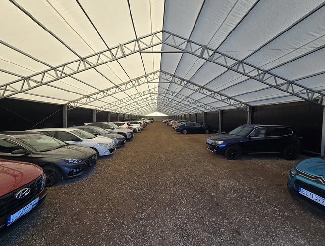 AleParking zadaszony - na parkingu stoją zaparkowane samochody. Teren jest kryty dachem. Samochody są ustawione w równych rzędach.