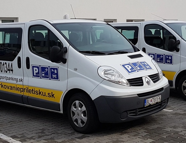 Dwa białe busy zaparkowane obok siebie na placu, na jednym z nich widoczny napis reklamowy z nazwą parkingu.