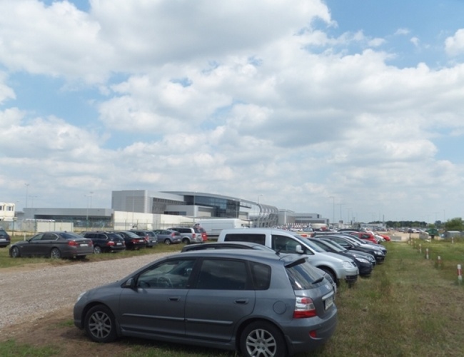 VALET Parking – En la hierba hay filas de coches aparcados. Al fondo se ve un edificio moderno. El cielo está parcialmente nublado.