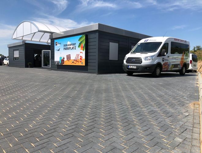 Tvoje parkovisko – Prostý, kamenistý dvor s kancelárskou budovou. Na parkovisku stojí biely minibus na prepravu.