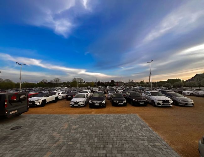 GoPark – Es stehen Reihen von Autos auf dem Parkplatz. Das umliegende Gelände ist offen und beleuchtet. Der Himmel ist teilweise bewölkt.