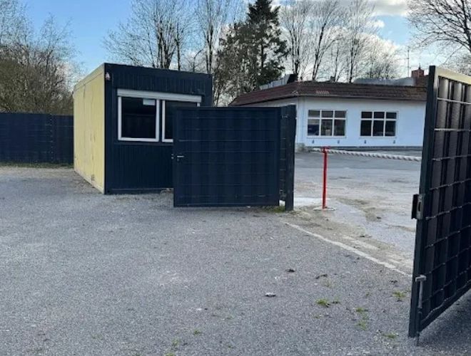 Parking Trust Park Fly – Wjazd na parking otoczony jest bramą. Znajduje się tuż przy ciemnym budynku.  Na końcu widać biały budynek.