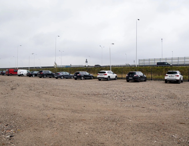 Jednoduché parkovanie štandard - Námestie s zaparkovanými autami. Pozemok nie je spevnený. Viditeľná je betónová bariéra a lampy.