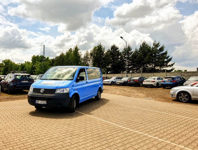 KTW Parking – En el área pavimentada hay un autobús azul. Al fondo, hay coches aparcados. El terreno está rodeado de árboles y una cerca.