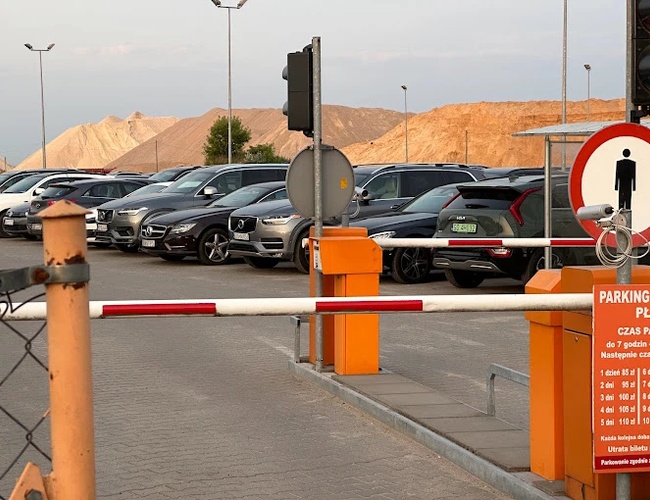 Oranžové parkovisko Modlin - Parkovisko je ohradené a osvetlené. Závorou sa riadi vjazd vozidiel. V pozadí sú vidieť kopce.