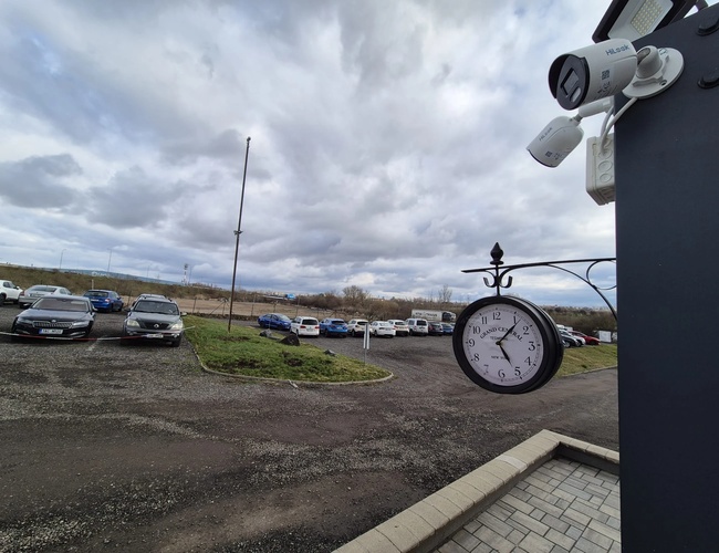 HostParking – Na parkovisku stoja početné autá. Pozemok je čiastočne oplotený. Vidieť je hodiny a kamery dohľadu.