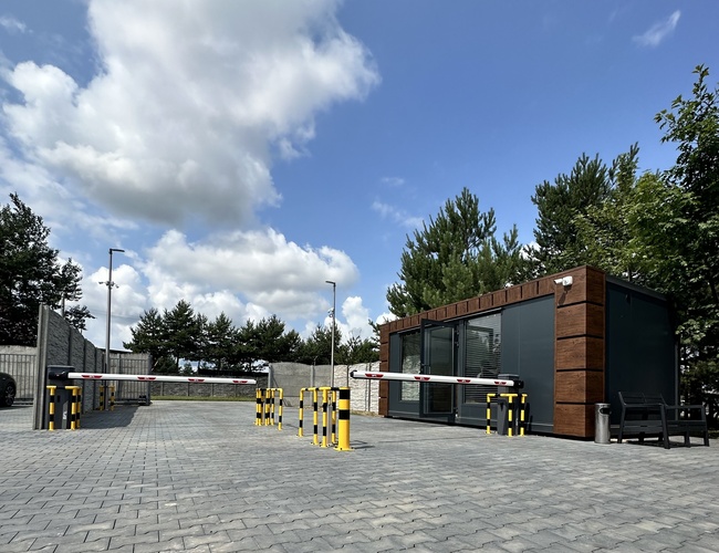 Empieza con nosotros - El parking está bien iluminado. Al lado se puede ver un pequeño edificio con fachada de madera. Hay una barrera en el recinto.