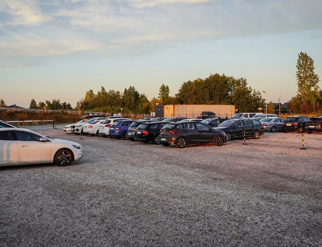 Parkovisko Sun - Parkovisko je štrkové a má veľa zaparkovaných áut. Okolie tvoria stromy a kontajnery. Na pozadí je vidieť oblohu s mrakmi.