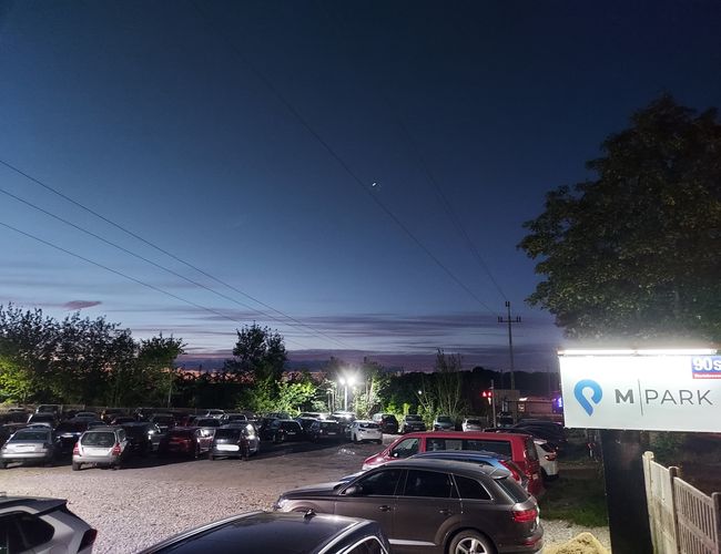 Aparcamiento MPark - La plaza está abierta y iluminada. Los coches están estacionados en varias filas. Al fondo se ven árboles y el cielo al anochecer.