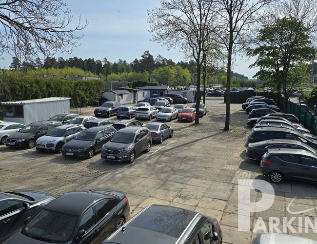 Parking Parkingos – Na zdjęciu widać otoczony zielenią parking. Różne samochody stoją w rzędach. Po lewej widoczny jest biały budynek.