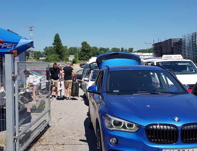 GOOD Parking - Parking jest ogrodzony i ochraniany. Samochody stoją w rzędzie, a ludzie pakują walizki. W tle widać niebo i drzewa.
