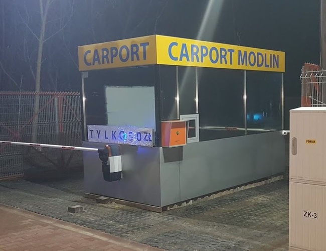 Parkovisko s prístreškom na auto – kontajner s nápisom, závorou a plotom. Parkovisko osvetlené v noci. Viditeľné kovové brány na ľavej strane.