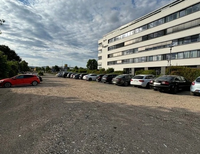 Špičkový parkovací personál - Parkovisko je nezpevnené a plné áut. Vedľa parkoviska stojí veľká biela budova. Obloha je čiastočne zamračená.