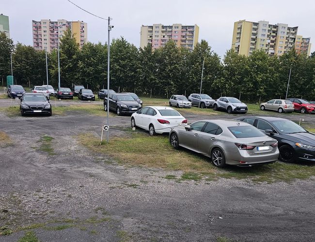 Estacionamiento Genial BZG - En la foto se pueden ver coches aparcados. El área está abierta y tiene una superficie de grava. Al fondo hay edificios residenciales.