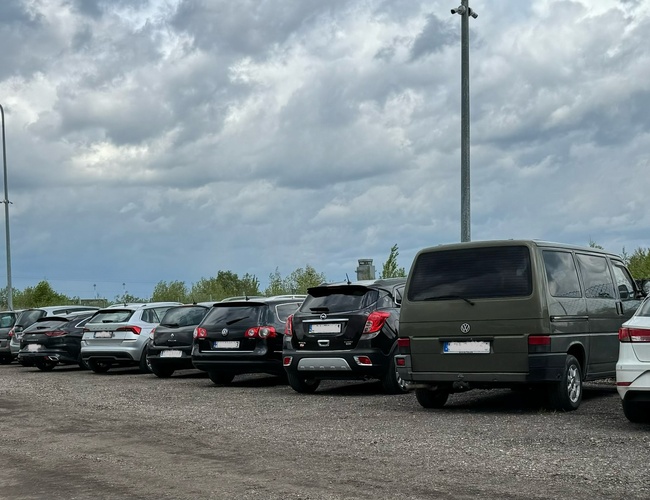Aparcamiento 66 - Una fila de coches está en la superficie de grava. Las farolas iluminan el área. Al fondo se pueden ver árboles y una valla.