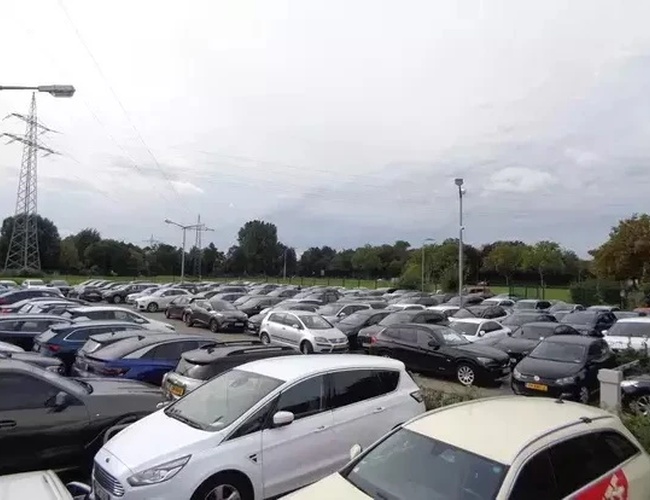 Parking Park&Fly Düsseldorf – wiele samochodów stoi obok siebie. Teren jest asfaltowy i otoczony zielenią. Oświetlenie jest zapewnione przez lampy.