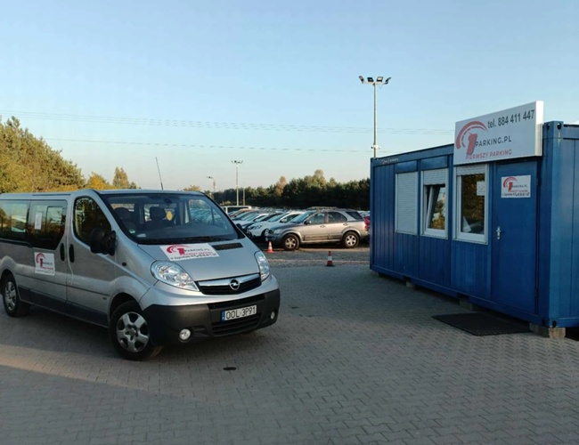 Primer estacionamiento - El minibús está frente al contenedor azul. En el fondo hay coches aparcados. El estacionamiento está pavimentado con adoquines.