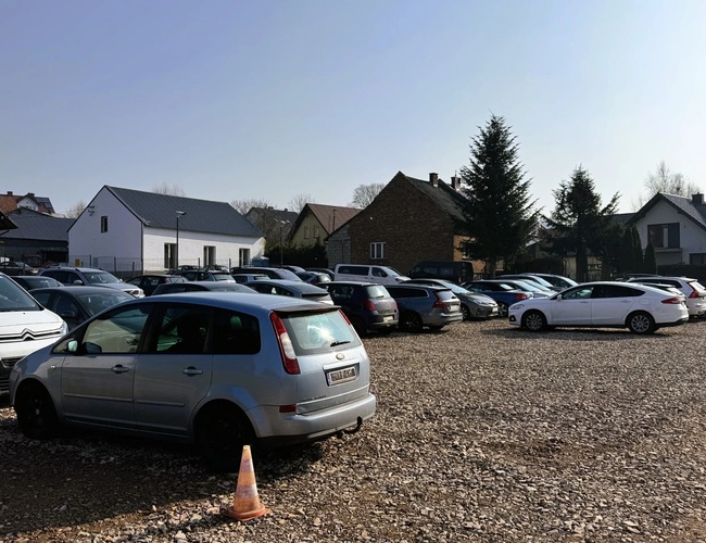 Parkovisko Amber - Na štrkovom pozemku sú zaparkované autá. Na pozadí sú viditeľné budovy a stromy.