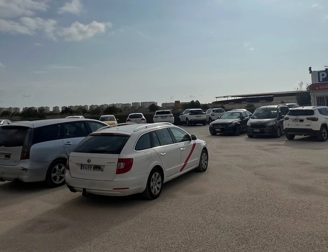 Parkeringsplass Punta Marina - Mange biler er parkert på plassen. I bakgrunnen ser man en bygning med bokstaven "P". På himmelen er det lette skyer.