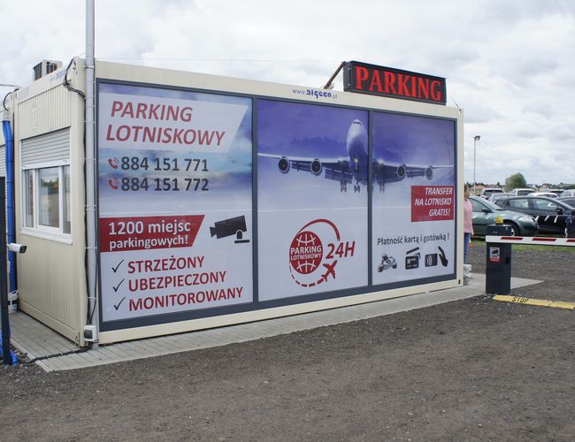 Parking Lotniskowy 24h - Kontener z reklamą będzie na pierwszym planie. Parking jest ogrodzony, a powierzchnia żwirowa. Na tle stoją samochody.