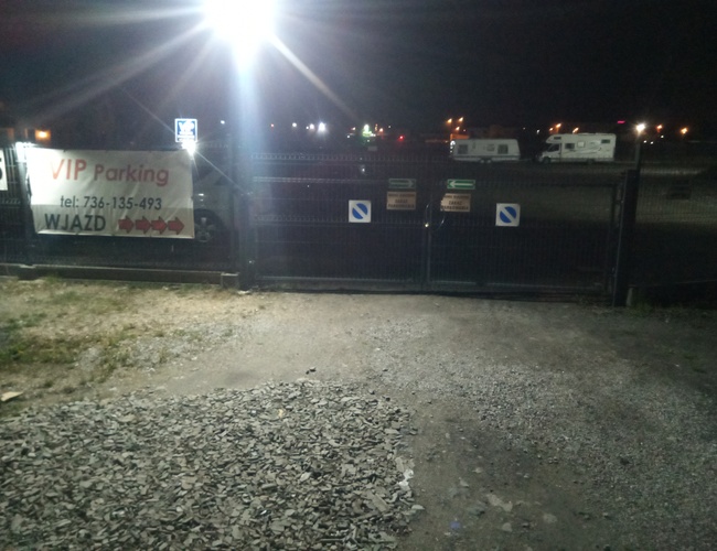 VIP Parking - Brama z napisem wjazd oświetlona nocą. W tle widoczne zaparkowane kampery. Teren jest ogrodzony.