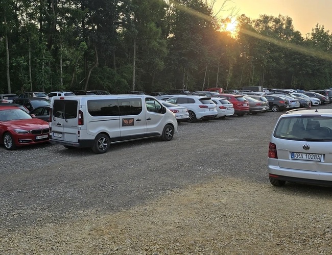Victory Cars Parking – Aparcamiento de grava lleno de coches. Sin vallas, el entorno son árboles. El sol se pone en el horizonte.