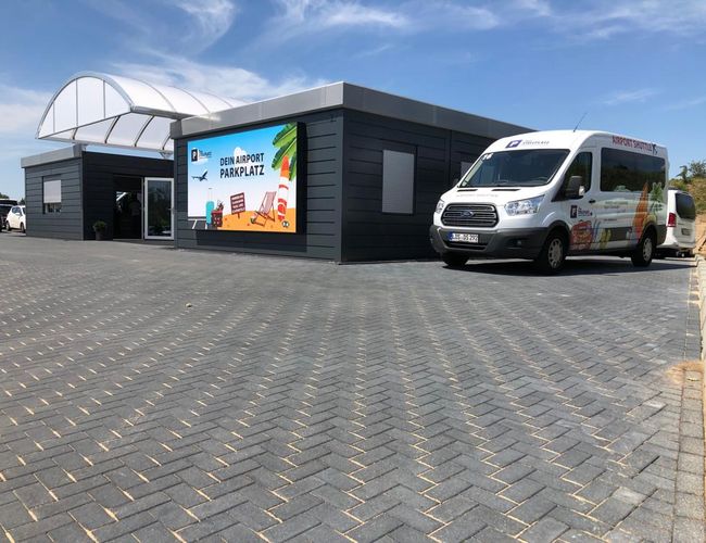 A parkolóhelyed – térkőből készült parkoló, modern épület szomszédságában van. Előtte egy fehér furgon áll reklámkampánnyal. Balra autók.