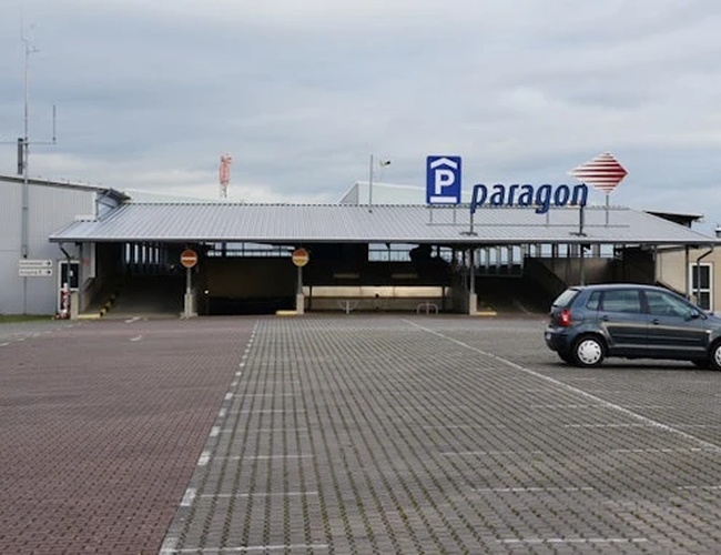 Widok na budynek z parkingiem przed wejściem. Na elewacji znajduje się oznakowanie parkingu, a przed budynkiem widoczne są miejsca postojowe i pojedyncze samochody.