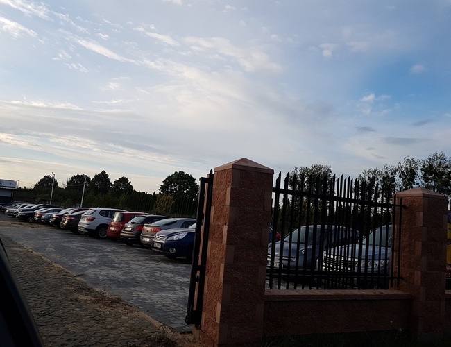 PARKING P24 PREMIUM - Parking jest ogrodzony wysokim metalowym płotem. Samochody stoją w rzędach na utwardzonej nawierzchni.