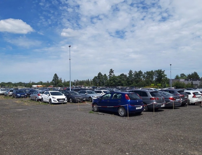 Parkovisko LOT 24 - Povrch je makadamový a otvorený. Na parkovisku stojí mnoho áut. V diaľke sú vidieť stromy.