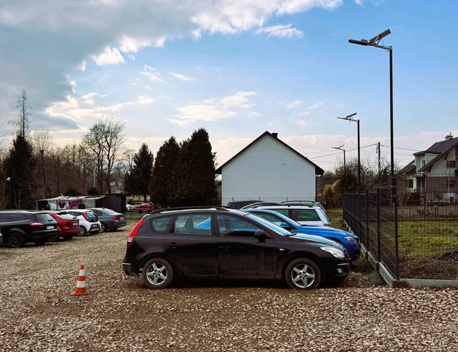 Parking Amber - Na placu stoi kilka samochodów. Teren jest ogrodzony i dobrze oświetlony latarniami. Obok parkingu rośnie kilka drzew.