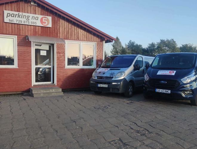 Parking na 5 – Przy drewnianym budynku stoją dwa samochody. Na ścianie widnieje duży szyld z numerem telefonu.