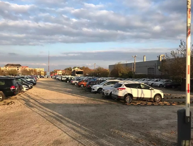 Żwirowy parking z dużą liczbą samochodów, szlabanem po prawej stronie i zabudowaniami w tle.