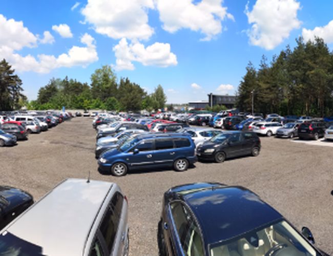 Parkplatz A1 – großes, offenes Gelände mit vielen Autos. Es ist von Bäumen und wenigen Zäunen umgeben. Der Untergrund ist kieselig.