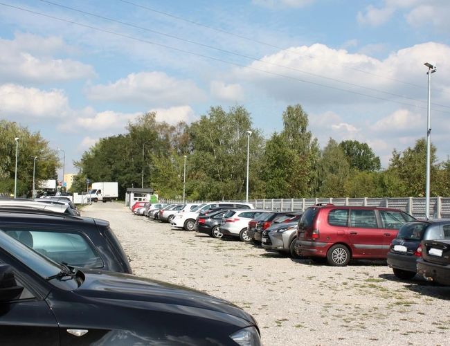 Parking Pienista 32 - Plac żwirowy z kilkoma samochodami. Teren jest otwarty i otoczony drzewami. Latarnie stoją przy krawędzi parkingu.