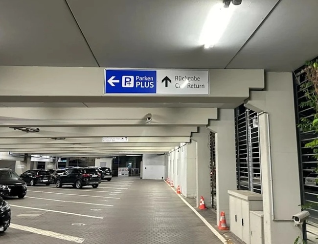 Wnętrze parkingu Parkenplus z szerokimi pasami ruchu, oznakowanymi miejscami postojowymi i tablicami kierunkowymi.