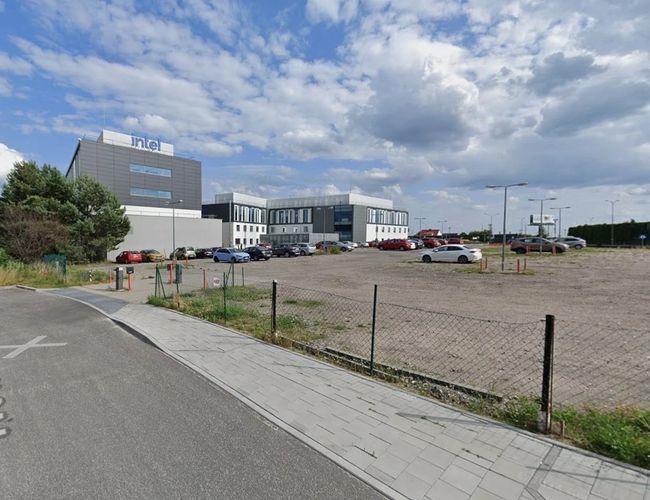 Parkplatz Piątka - unbefestigter Parkplatz mit Netz, in der Nähe des Bürogebäudes. Die Straße mit Gehweg führt zum Eingang. In der Ferne sind Autos unter dem Gebäude zu sehen.