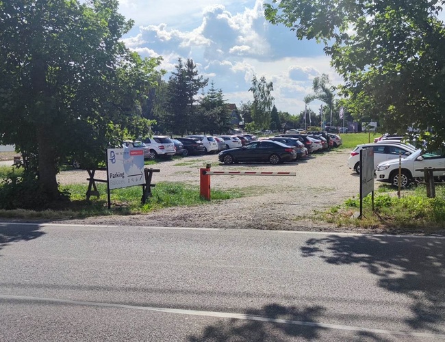Parking Guardia - Teren jest ogrodzony i ma szlaban. Widoczne są zaparkowane samochody i budka strażnika. Drzewa dają cień.