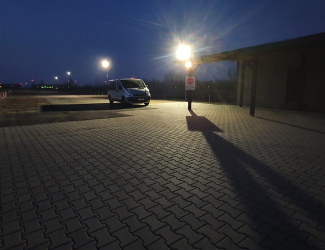 WIZ-PARK - Aparcamiento de adoquines iluminado con farolas. Hay un coche aparcado. El edificio se encuentra a la derecha.